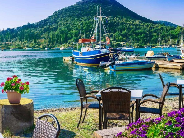 Inviting café seating by the tranquil waters of Lefkada, with colourful boats moored and lush hills in the background.