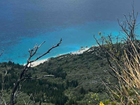 Aerial view of a coastal land area in Kalamitsi, showcasing the lush greenery, the clear turquoise waters of the Ionian Sea, and the sandy beach with visible structures in the distance.